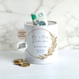May include: White ceramic money box with a gold circle around the text "Mr & Mrs Richardson" and the words "Wedding Fund Engaged 02.02.23". The design features a watercolor-style illustration of dried grasses in shades of beige and gold.