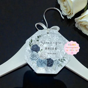 May include: A personalised wedding hanger tag with the name "Jennifer" and the word "BRIDE" printed on it. The tag features a floral design with blue and white roses and the date 03.06.2026. The tag is attached to a white hanger.