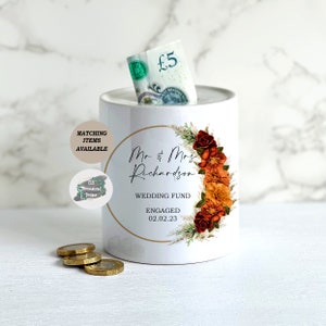 May include: White ceramic money box with a floral design in shades of orange and red. The text "Mr & Mrs Richardson" is printed on the front, along with "Wedding Fund" and "Engaged 02.02.23".