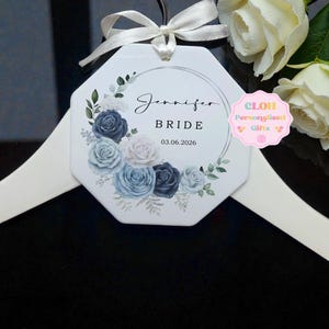 May include: White octagonal hanger tag with a floral design in shades of blue and white. The tag reads "Jennifer BRIDE 03.06.2026". A white ribbon is tied at the top. The tag is hanging on a white hanger.