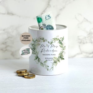 May include: White ceramic money box with a green floral design and the text "Mr & Mrs Richardson Wedding Fund Engaged 02.02.23".
