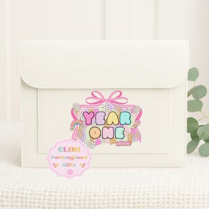 May include: Cream-coloured rectangular clutch bag with a flap closure. The front displays a colourful graphic with the words "YEAR ONE", a rainbow, and flowers. A pink sticker with text is on the lower left.
