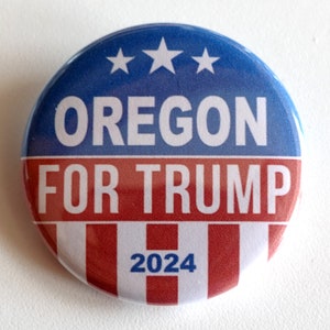 May include: A round button with a blue and red design. The top half is blue with three white stars, and the bottom half is red and white striped. The text "OREGON FOR TRUMP 2024" is printed in white.