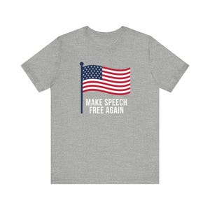 Puede incluir: Camiseta gris con un diseño gráfico de la bandera estadounidense y el texto blanco "MAKE SPEECH FREE AGAIN". La bandera es roja, blanca y azul.