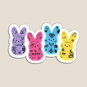 Peut inclure: Trois Peeps en guimauve de couleur pastel en forme de lapin. Chaque Peep est recouvert de tatouages noirs et blancs, y compris des cœurs, des étoiles, des crânes et des mots comme "crybaby", "love" et "daddy".