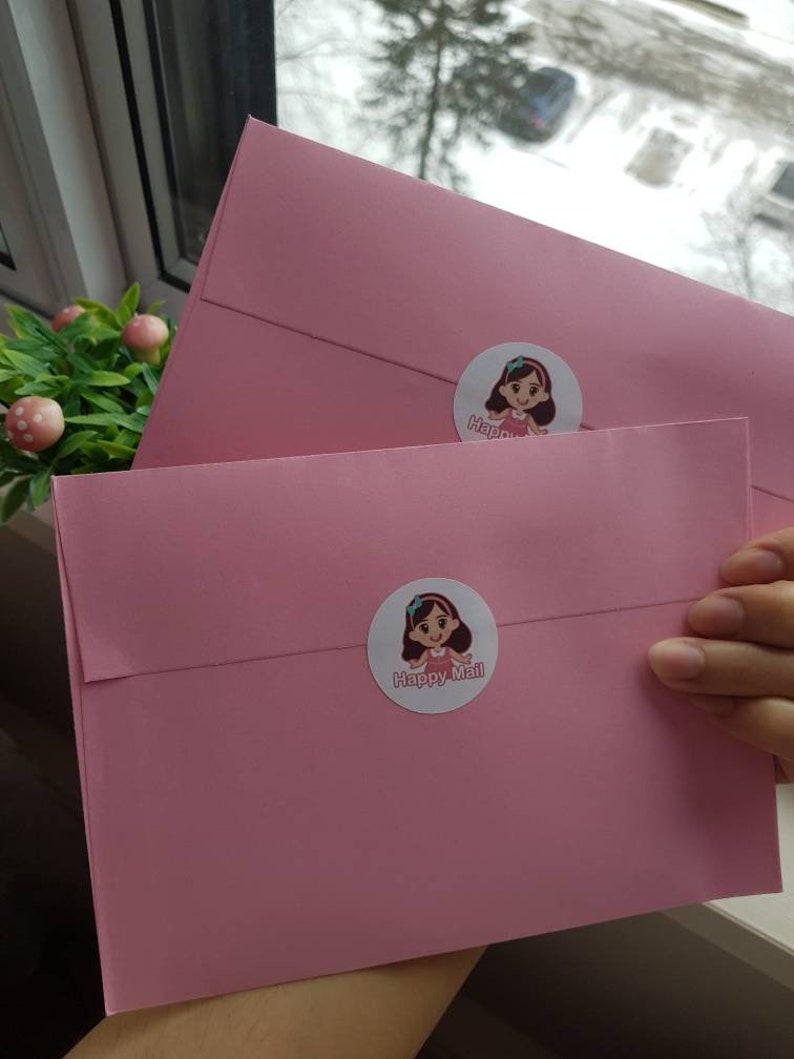 May include: Two pink envelopes with a cartoon girl sticker on each. The sticker on the top envelope says "Happy" and the sticker on the bottom envelope says "Happy Mail".