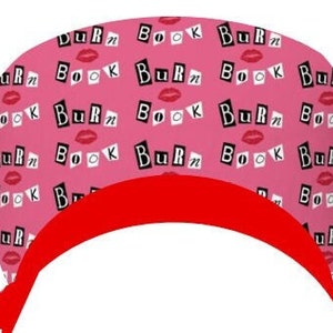May include: A pink scrub cap featuring a repeating pattern of the text "Burn Book" in a ransom-note style font, interspersed with small red lip icons. The cap has a solid red fabric band along the bottom edge with ties extending from the sides.