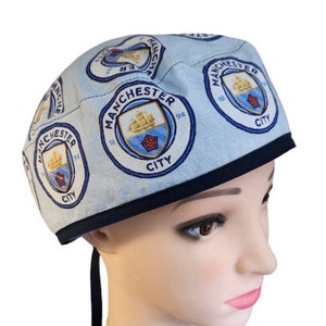 May include: A light blue scrub hat with a navy blue trim and a repeating pattern of the Manchester City Football Club logo. The logo features a ship, a rose, and the text "Manchester City 1894".