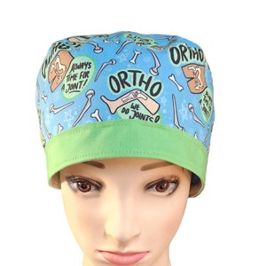May include: A blue surgical cap with a green band. The cap is patterned with bones, joints, and the word "ORTHO", along with the phrases "ALWAYS TIME FOR A JOINT!" and "WE DO JOINTSO".