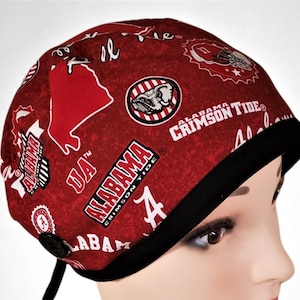 May include: Red and black scrub hat with a button and ties. The hat is covered in a print of the Alabama Crimson Tide logo, the state of Alabama, and the letters "UA".