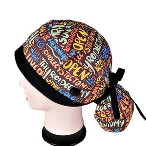 May include: A brown scrub cap with a black brim and a black tie closure. The cap is covered in colorful text, including words like "OPEN", "HERNIA", and "THYROIDECTOMY". A black button is on the side.
