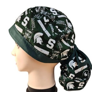 May include: A green and white patterned scrub cap featuring the Michigan State Spartans logo and text. The cap has a dark green brim and a matching tie in the back. The design includes the letters "S", "MSU", and "Spartans".