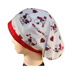 May include: A white and red patterned cap featuring Snoopy characters and red hearts. The cap has a red trim and is made of fabric. The design includes Snoopy holding cards and hearts, perfect for a themed gift.