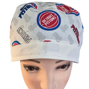 May include: A white scrub cap featuring a repeating pattern of the Detroit Pistons basketball team logo in red, white, and blue, along with the team name in black and blue text, finished with a solid white folded band at the bottom edge.