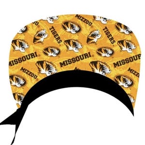 May include: A scrub cap featuring a repeating pattern of Missouri Tigers logos and the text MIZZOU and MISSOURI in black on a gold background. The cap has a solid black band along the bottom edge and black fabric ties extending from the sides.