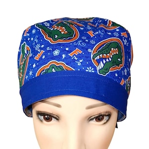 May include: A blue scrub cap featuring a repeating pattern of green alligator heads with orange outlines and the orange letter F. The fabric includes small white floral and geometric accents against a blue background with a solid blue band.