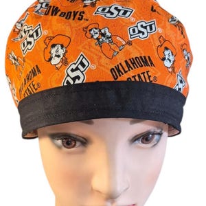 Könnte beinhalten: Eine orangefarbene OP-Haube mit einem sich wiederholenden Muster aus dem Logo der Oklahoma State University, dem Schriftzug Cowboys und dem Maskottchen Pistol Pete. Die Haube hat ein einfarbiges schwarzes Stoffband am unteren Rand.