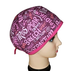 May include: A pink and purple surgical scrub hat with a pink trim. The hat is covered in white text that says "TRUSAPGAR", "TONS BREECH BABY!!", "CERVIX", "POSTPARTUM", "♡B/ABOR C-SECTION", "R", "0.0.0", "TENS", "♡B", "TS!!".
