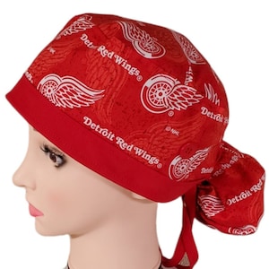May include: Red and white surgical scrub hat with a Detroit Red Wings logo pattern. The hat has a button on the top and a tie at the back.