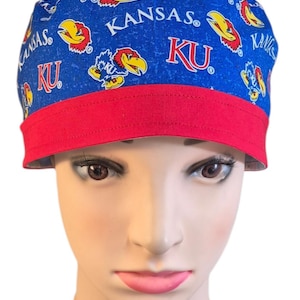 May include: A blue scrub cap featuring a repeating pattern of the Kansas Jayhawk mascot and the text KU and KANSAS. The cap has a solid red band along the bottom edge. The fabric has a textured blue background with white and red graphic elements.