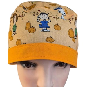 May include: A beige scrub cap featuring a repeating pattern of cartoon characters, pumpkins, and autumn leaves. The cap has a bright orange band at the base. The characters include a girl in a blue dress and Snoopy.