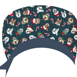 Könnte beinhalten: Eine dunkelblaue OP-Haube mit einem Muster aus verschiedenen Hunderassen, die Weihnachtsmützen und Weihnachtspullover tragen. Auch Zuckerstangen sind auf dem Stoff verstreut. Die Haube hat ein dunkelblaues Band.
