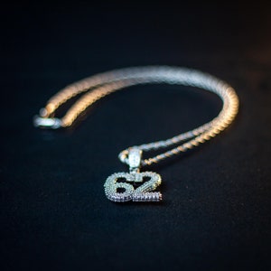 Puede incluir: Un collar de cadena de plata con un gran colgante en forma de n&uacute;mero 62 adornado con diamantes.
