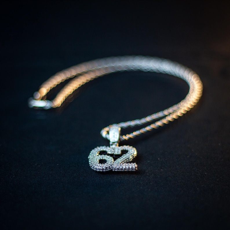 Puede incluir: Un collar de cadena de plata con un gran colgante en forma de n&uacute;mero 62 adornado con diamantes.