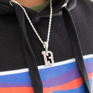 Puede incluir: Un collar de cadena de cuerda plateada con un gran colgante plateado en forma del n&uacute;mero "13". El colgante est&aacute; cubierto de peque&ntilde;os diamantes de imitaci&oacute;n transparentes.