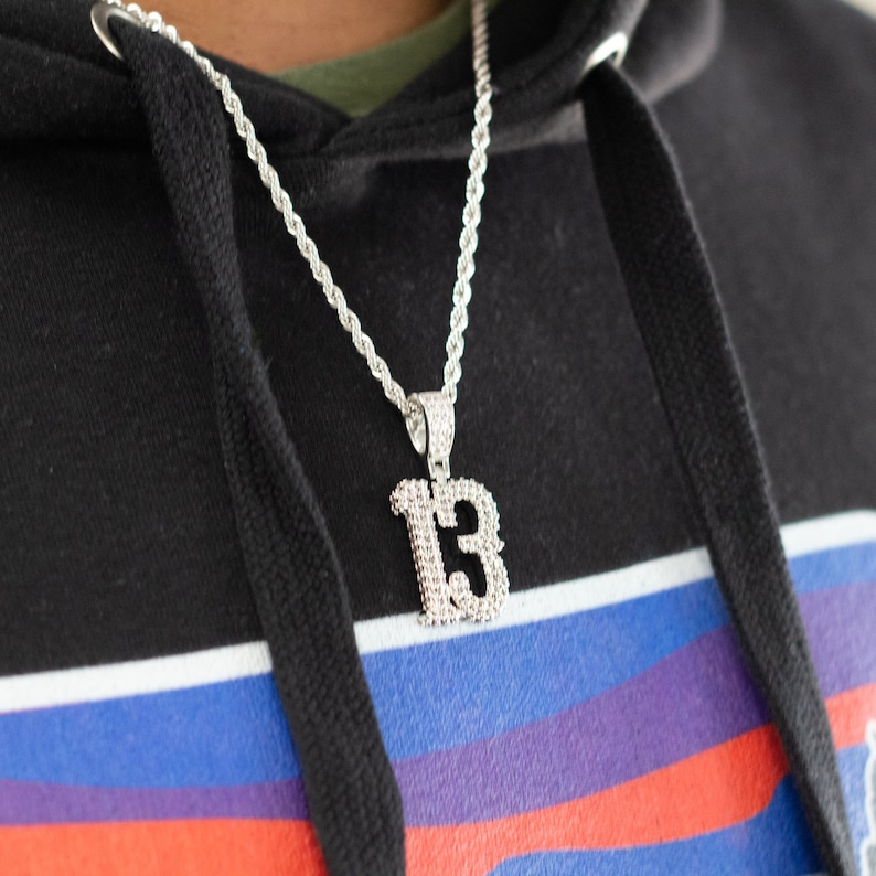 Puede incluir: Un collar de cadena de cuerda plateada con un gran colgante plateado en forma del n&uacute;mero "13". El colgante est&aacute; cubierto de peque&ntilde;os diamantes de imitaci&oacute;n transparentes.