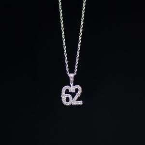 Puede incluir: Un collar de cadena de plata con un colgante engastado con diamantes que presenta el n&uacute;mero "62".