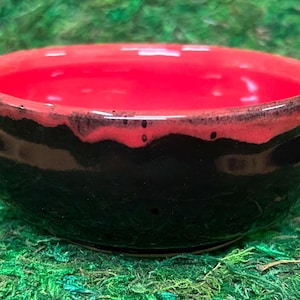 May include: A small ceramic bowl with a glossy finish. The exterior is a deep, solid black, while the interior and the upper rim feature a vibrant, bright red glaze that drips slightly over the black surface in a textured, uneven pattern.