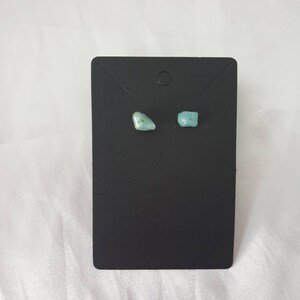 May include: A pair of small, light blue gemstone earrings on a black display card.