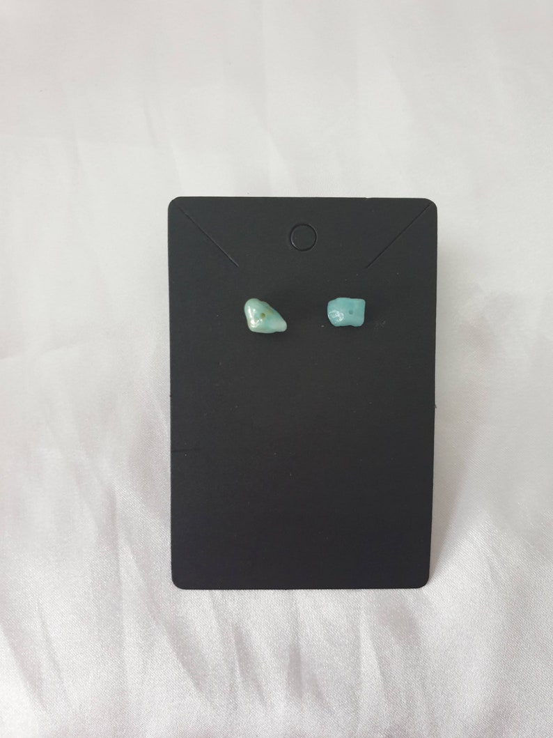 May include: A pair of small, light blue gemstone earrings on a black display card.