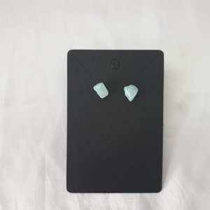 May include: A pair of small, light blue gemstone earrings on a black display card.