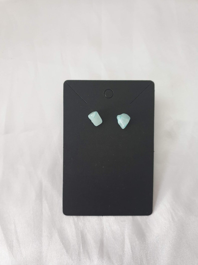 May include: A pair of small, light blue gemstone earrings on a black display card.