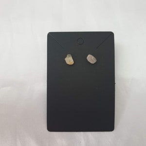 May include: A pair of small, raw crystal earrings on a black display card. The earrings are a light brown and a light gray.