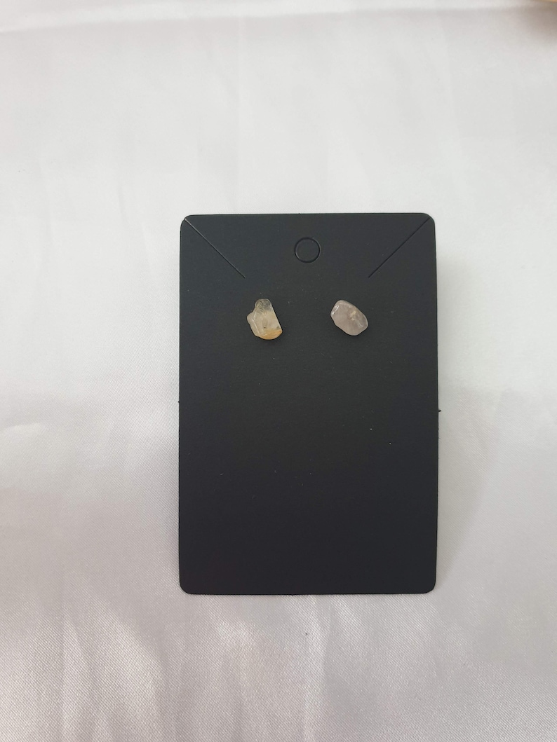 May include: A pair of small, raw crystal earrings on a black display card. The earrings are a light brown and a light gray.