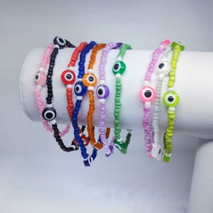 May include: A set of colorful beaded bracelets with evil eye charms. The bracelets are made with small, colorful beads and feature a variety of colors, including pink, orange, blue, green, and white. Each bracelet has a single evil eye charm in the center.