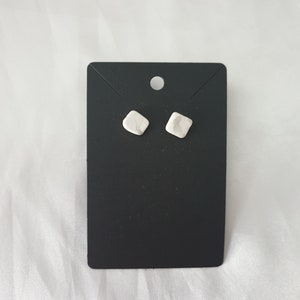 May include: A pair of small, white, square-shaped earrings on a black display card.