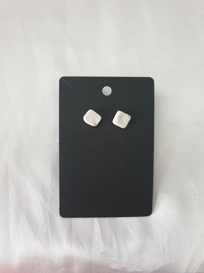 May include: A pair of small, white, square-shaped earrings on a black display card.