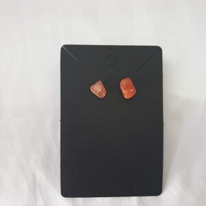 May include: A pair of small, orange-red gemstone earrings on a black display card.