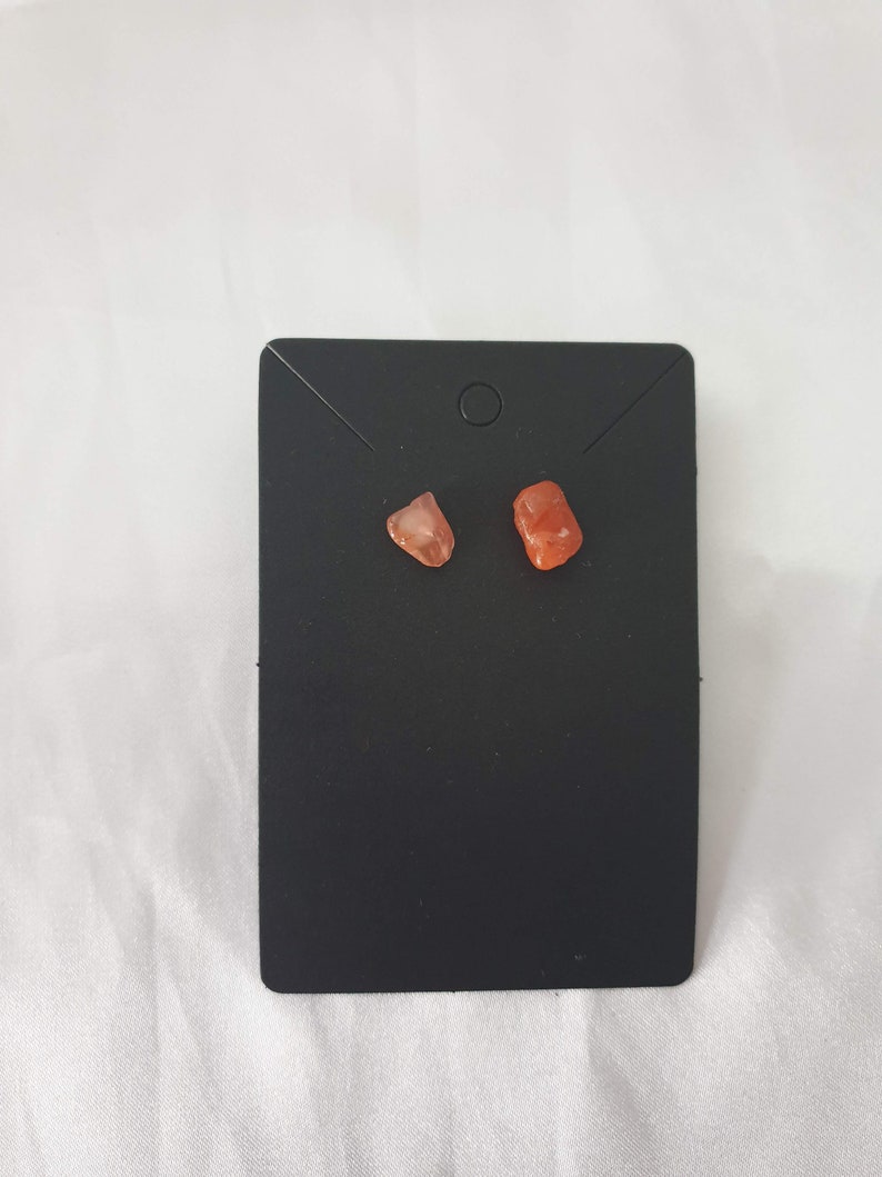 May include: A pair of small, orange-red gemstone earrings on a black display card.