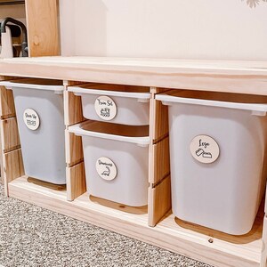 Pu&ograve; includere: Un'unit&agrave; di stoccaggio in legno con quattro contenitori bianchi. I contenitori sono etichettati con cerchi di legno che dicono "Dress Up", "Train Set", "Dinosaurs" e "Lego".