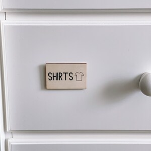 May include: A white drawer with a wooden label that says "SHIRTS" with a simple line drawing of a t-shirt.