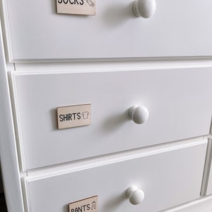 May include: White dresser with three drawers. Each drawer has a wooden label with black text and a small icon. The top drawer label says "SOCKS" with a sock icon. The middle drawer label says "SHIRTS" with a shirt icon. The bottom drawer label says "PANTS" with a pants icon.