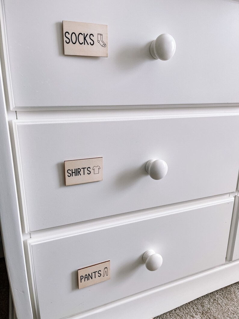 May include: White dresser with three drawers. Each drawer has a wooden label with black text and a small icon. The top drawer label says "SOCKS" with a sock icon. The middle drawer label says "SHIRTS" with a shirt icon. The bottom drawer label says "PANTS" with a pants icon.