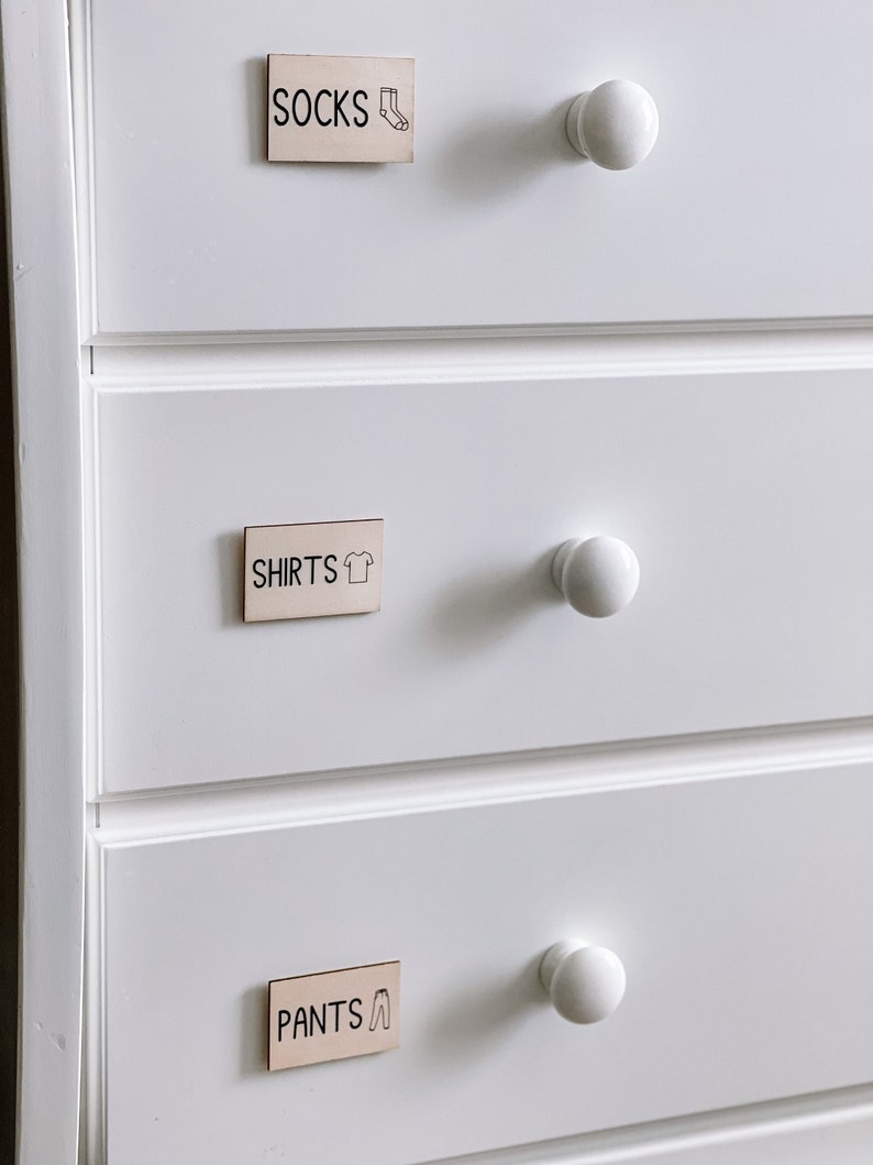 May include: White dresser with three drawers. Each drawer has a wooden label with a different category of clothing: "Socks", "Shirts", and "Pants". Each label has a small icon representing the clothing category.