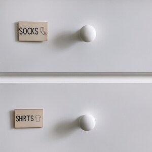 May include: Two white drawers with wooden knobs. The top drawer has a label that says "SOCKS" with a sock icon. The bottom drawer has a label that says "SHIRTS" with a t-shirt icon.