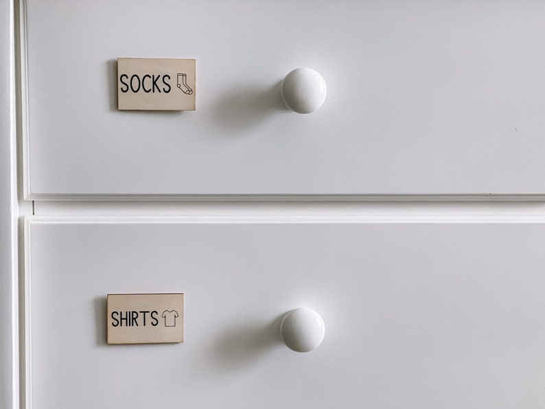 May include: Two white drawers with wooden knobs. The top drawer has a label that says "SOCKS" with a sock icon. The bottom drawer has a label that says "SHIRTS" with a t-shirt icon.